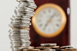 A close up of a tall stack of coins, and a couple of them scattered beside the stack. The rest of the picture is blurred, however, we can see a big clock on the wall in the distance.